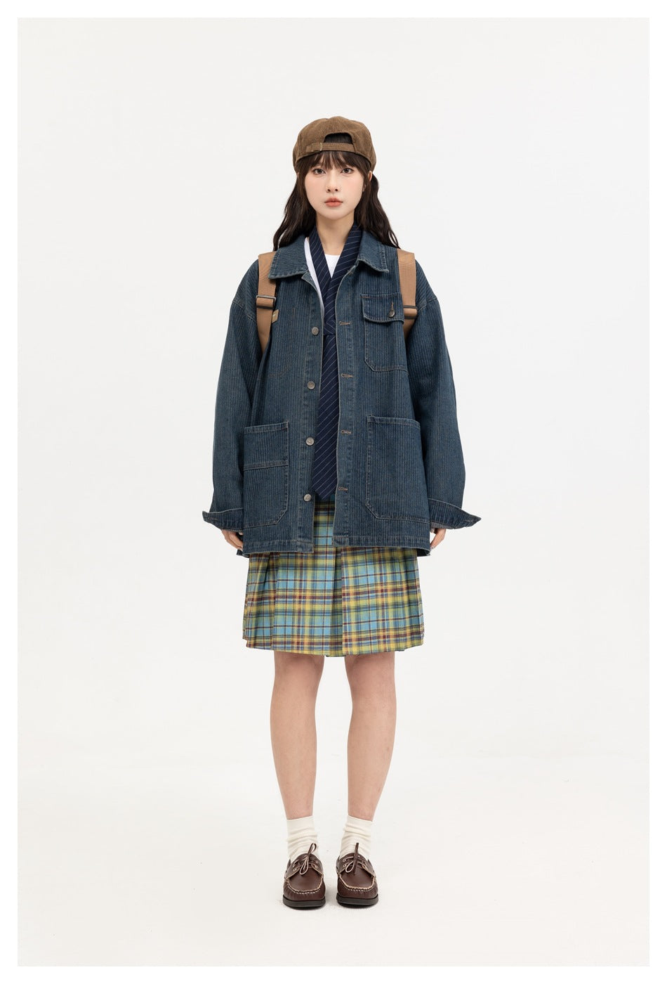 Model wearing an oversized dark blue denim jacket open over a striped tie, plaid pleated skirt, cap and backpack, front view