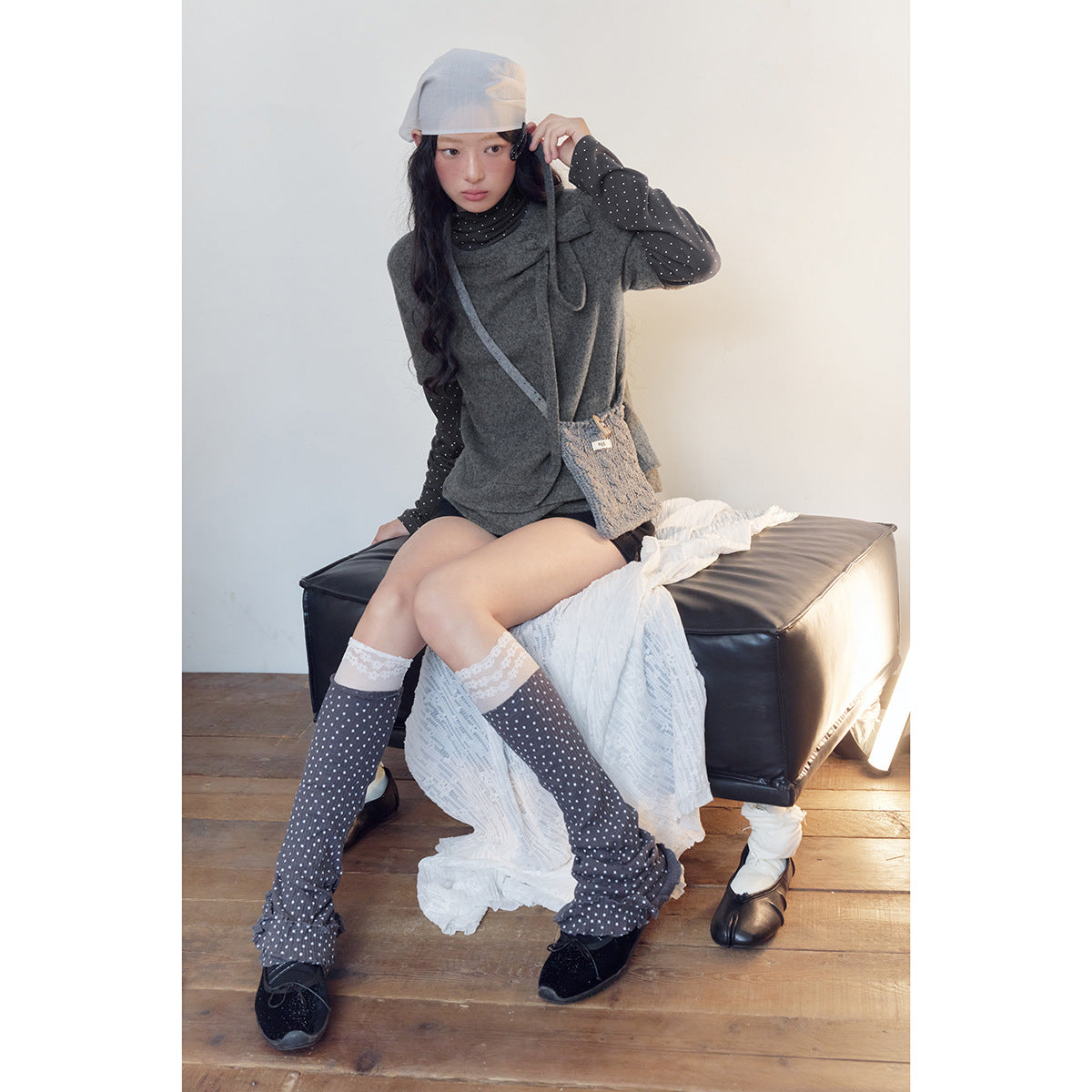 Seated model wearing a charcoal gray bow-detail top over a black polka-dot layer, styled with matching dotted leg warmers.