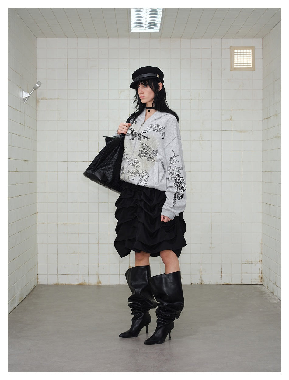 Model wearing a gray zip-up hoodie with black illustrated graphics, a ruffled black skirt, and slouchy black boots.
