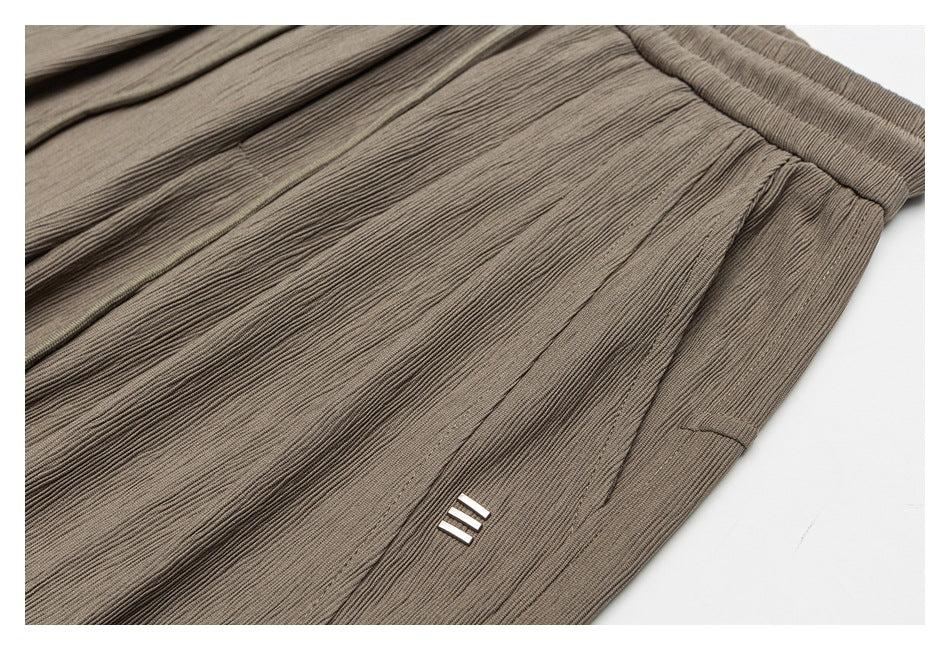 Close-up of taupe pants showing a side pocket, pleats, and a small three-bar detail.