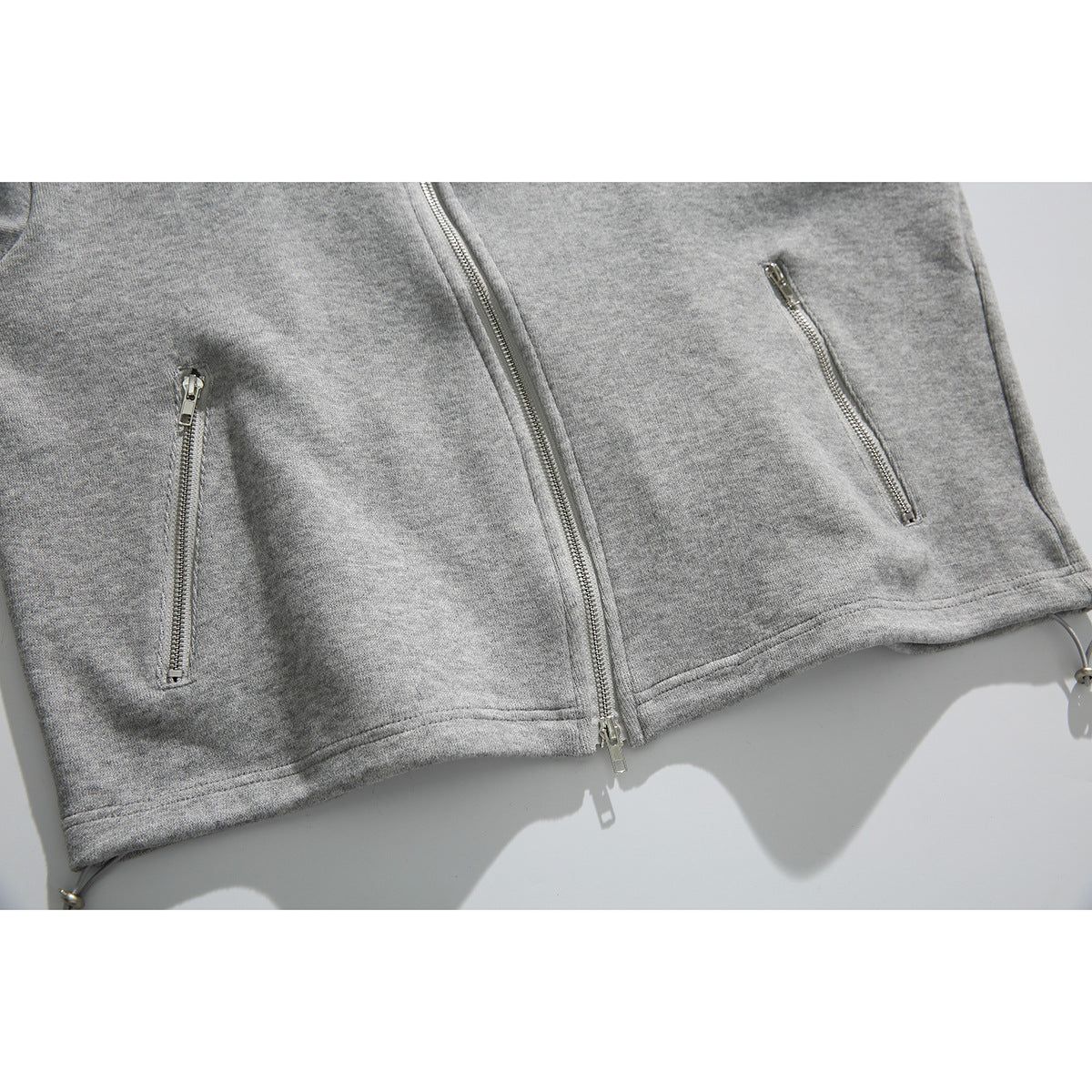 Close-up of a gray zip-up hoodie with a center zipper, two vertical zip pockets, and hem drawcords.