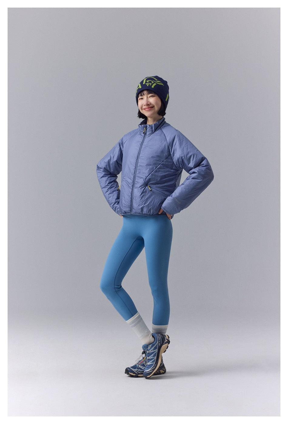 Model wearing a periwinkle-blue padded zip jacket and blue leggings, with a navy beanie and trail shoes.