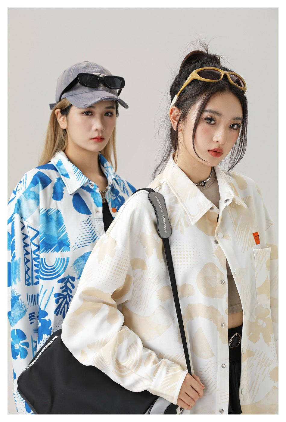 Two models wearing oversized printed shirts in blue and cream, close-up with sunglasses, cap and black shoulder bag