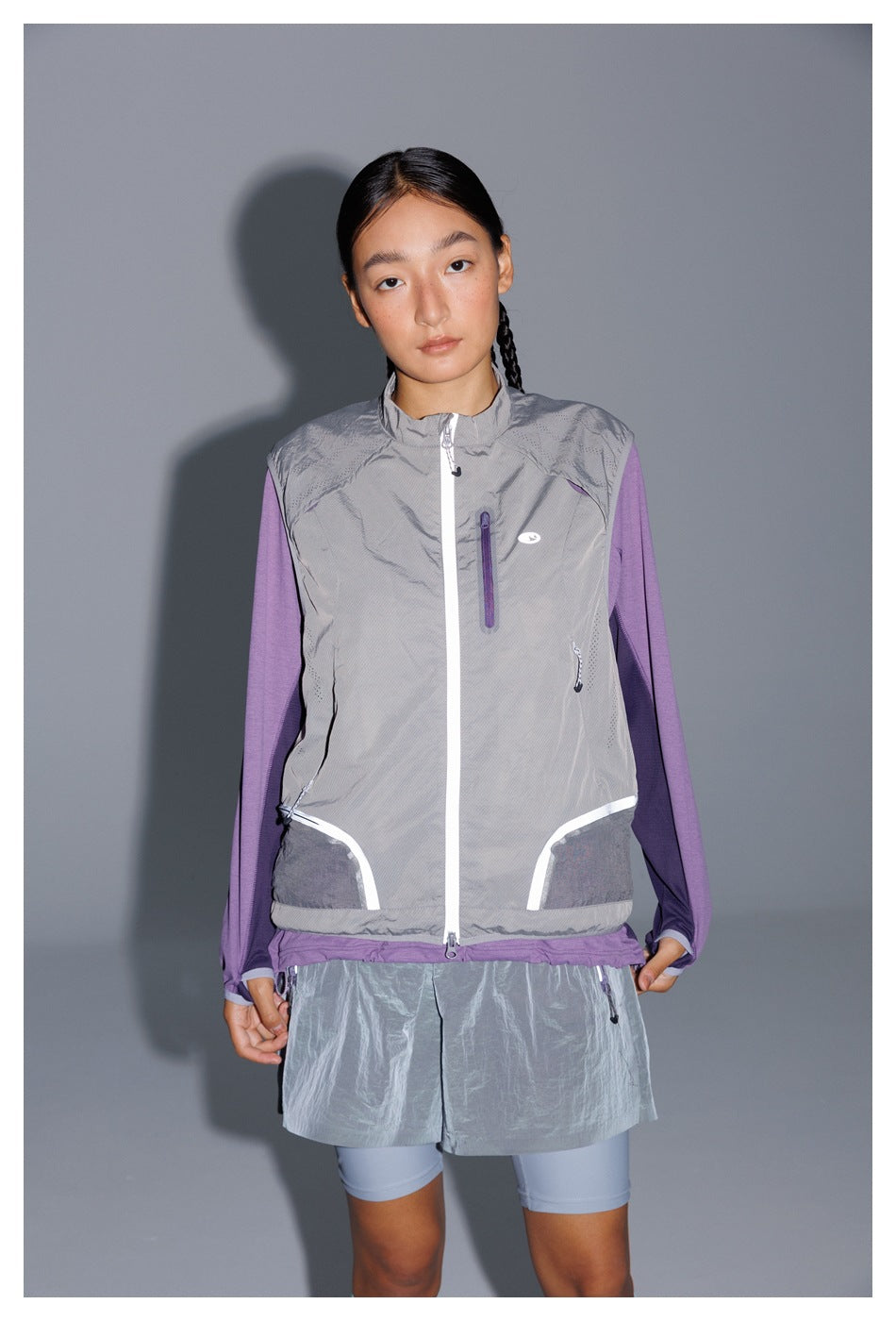 Front view of a model wearing a gray zip vest with white trim over a purple long-sleeve top and gray shorts.