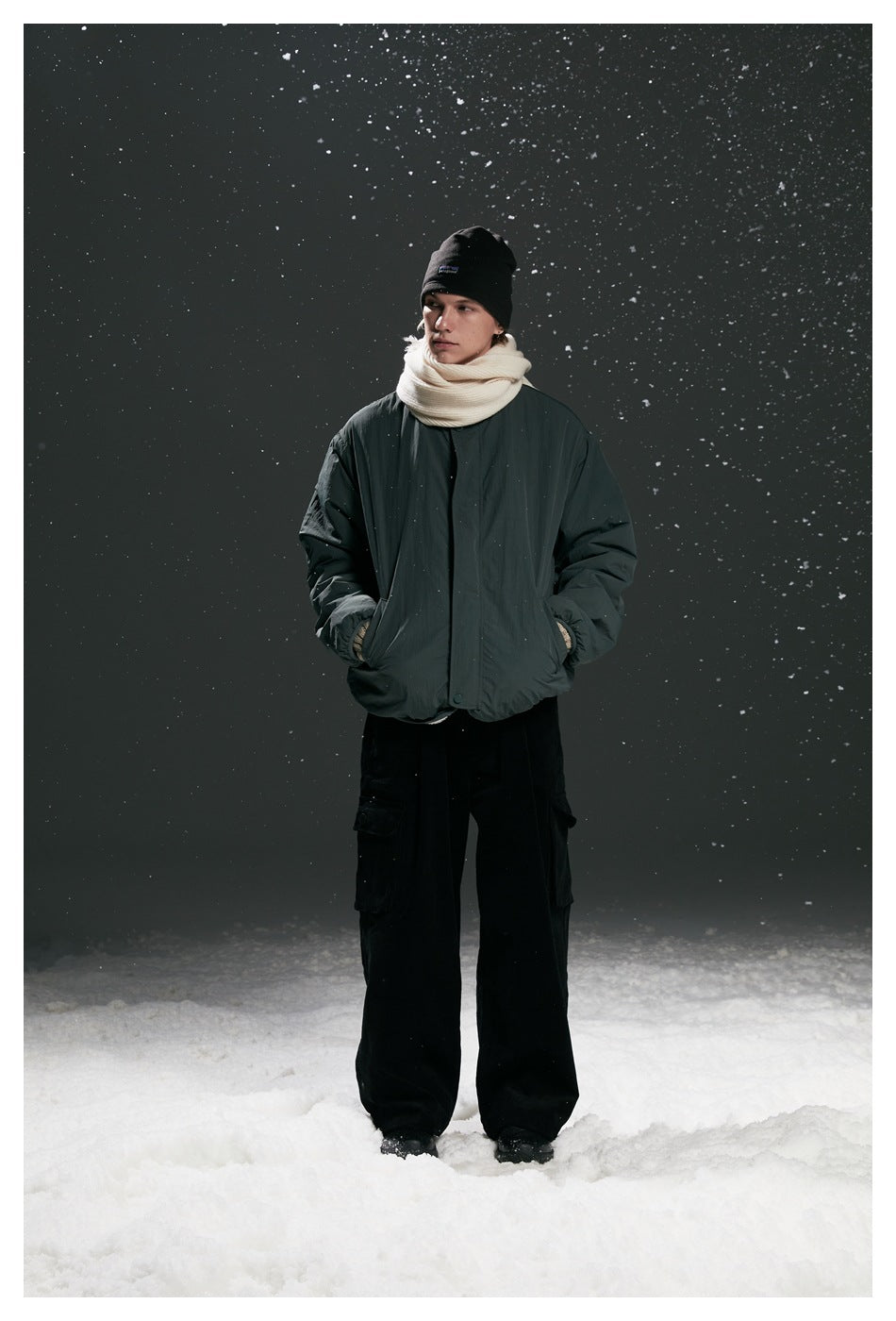 Model wearing a dark charcoal padded jacket, black beanie, cream scarf, and loose black cargo pants in falling snow.