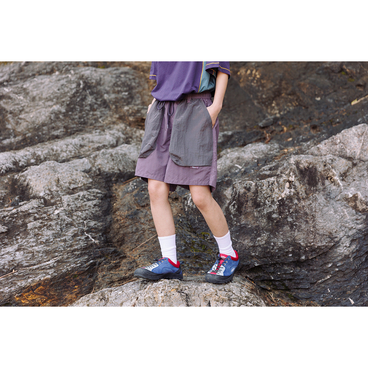 Model outdoors wearing muted purple shorts with charcoal front pockets, shown against a rocky background.