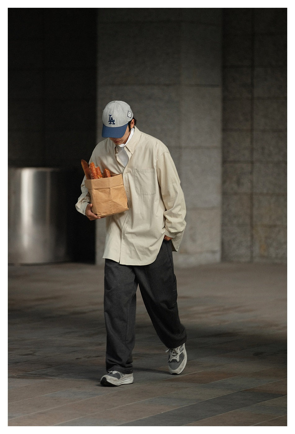 Man walking in a cream oversized button-down shirt with chest pocket, dark pants and a cap, carrying a paper bag