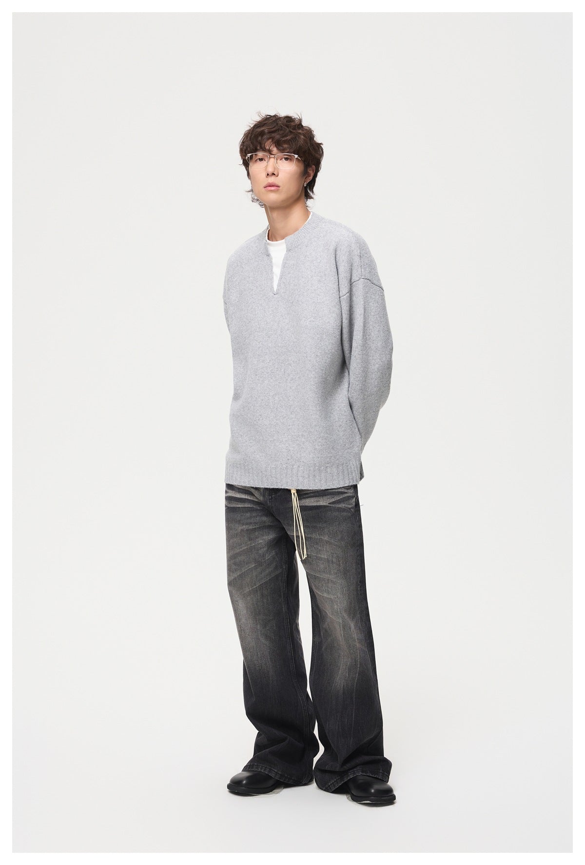 Man standing in a light gray knit sweater with white layer at the neckline and faded black wide-leg jeans