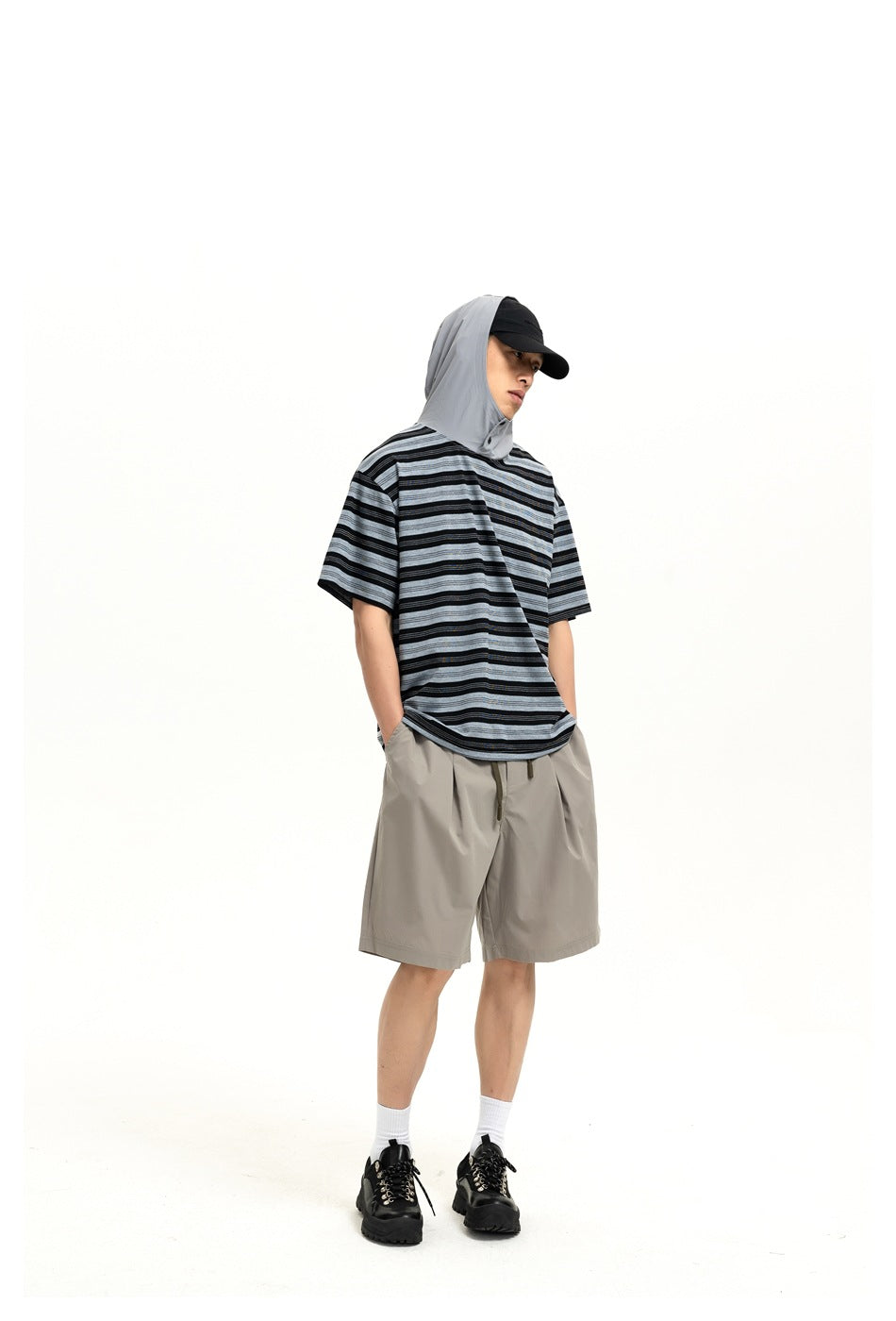 Model wearing gray wide-leg pleated shorts with a striped hooded tee, cap and black shoes, front view