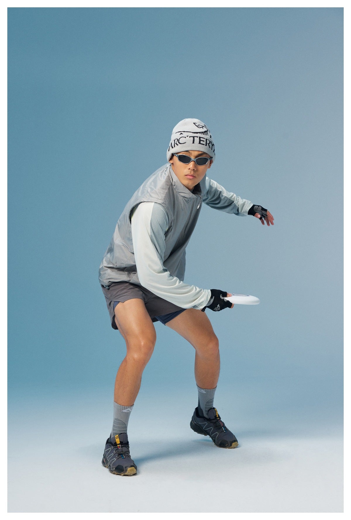 Man in an athletic pose wearing a light gray sleeveless vest over a pale long-sleeve top and gray shorts.
