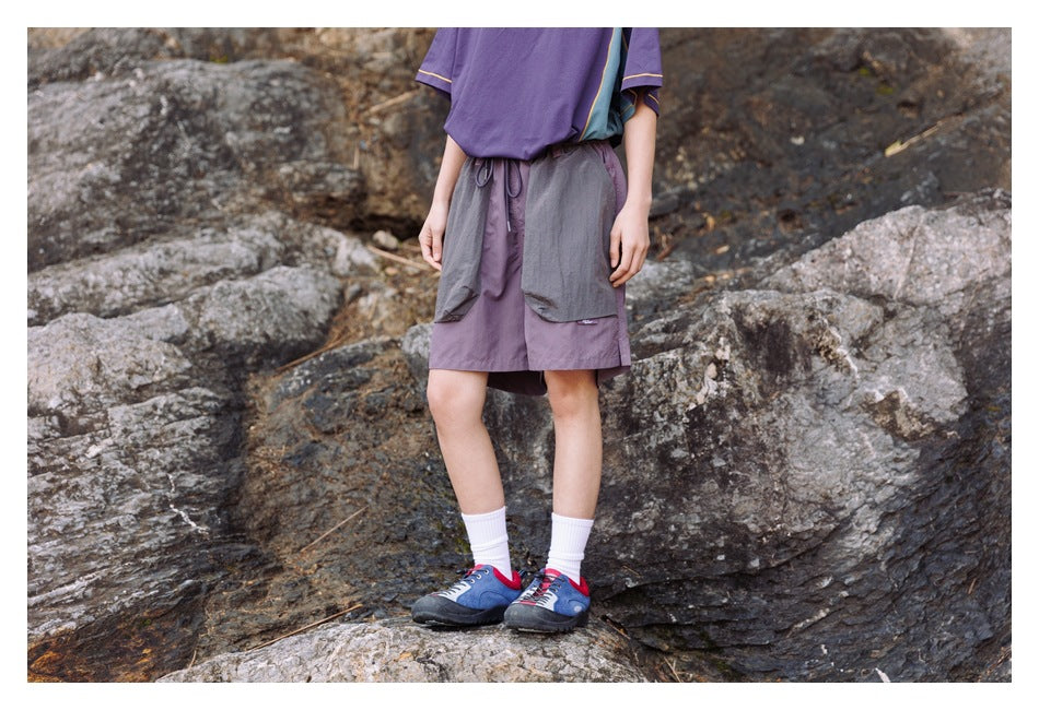 Person wearing purple drawstring shorts with gray patch pockets, standing on rocks, cropped below the chest