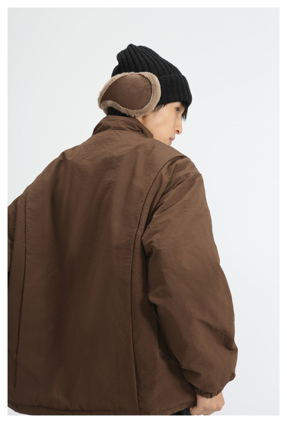 Rear view of a model wearing a brown jacket with a high collar and loose sleeves, paired with a black knit earflap hat.