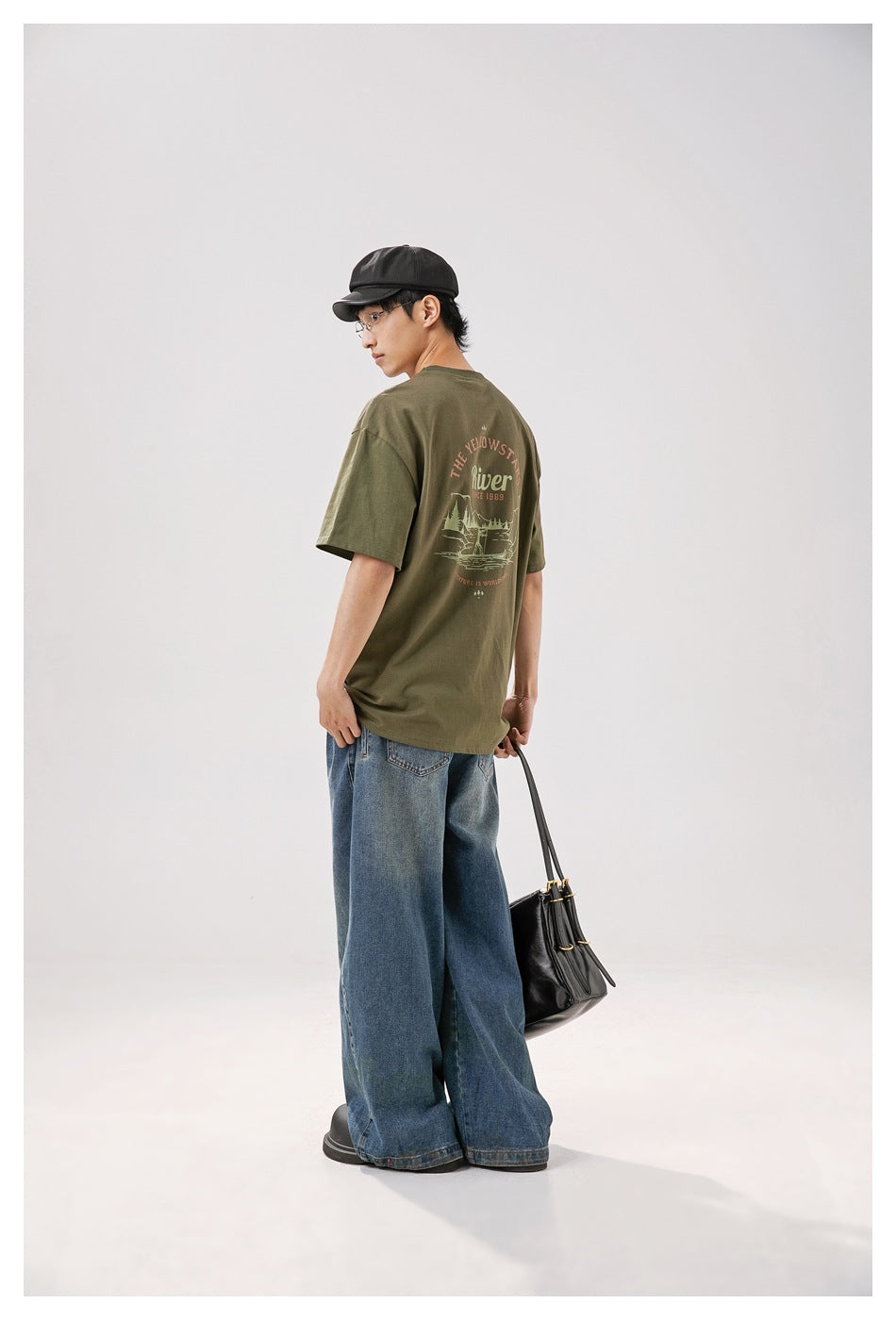 Model in an olive T-shirt and wide-leg jeans, showing the large illustrated graphic on the back.
