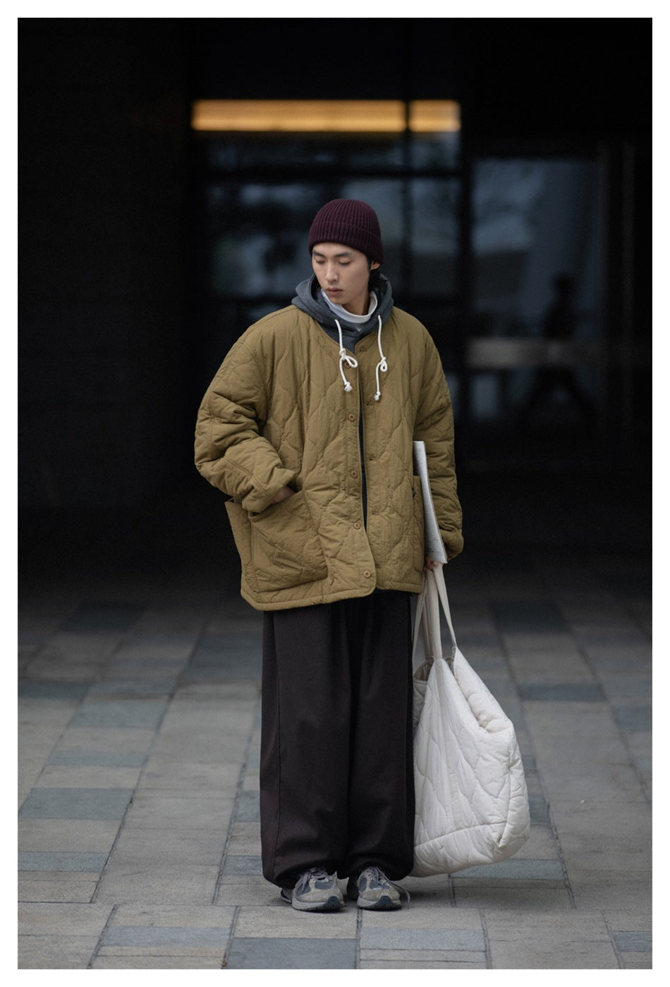 A person models an olive quilted button-front jacket with large patch pockets, layered over a gray hoodie.