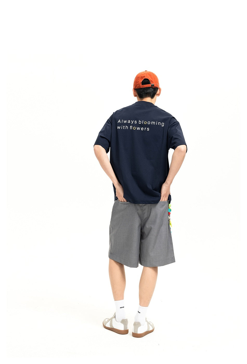 Rear view of a model in a navy T-shirt printed “Always blooming with flowers,” gray shorts, and an orange cap.