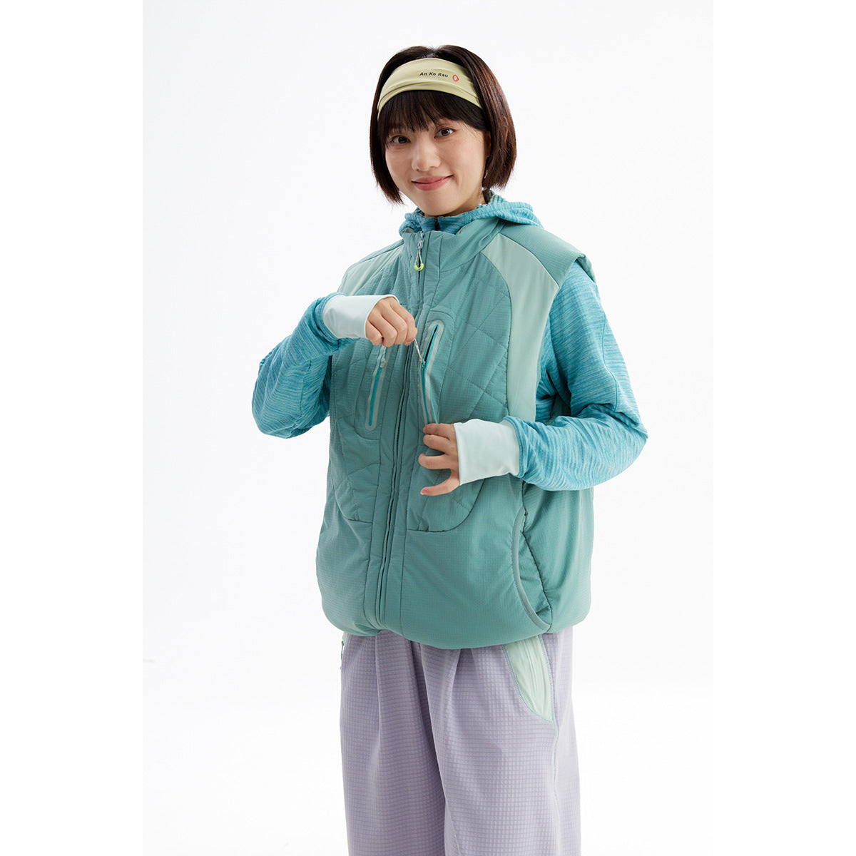 Model in a teal quilted sleeveless vest over a blue hooded top, showing the front pockets and zipper.