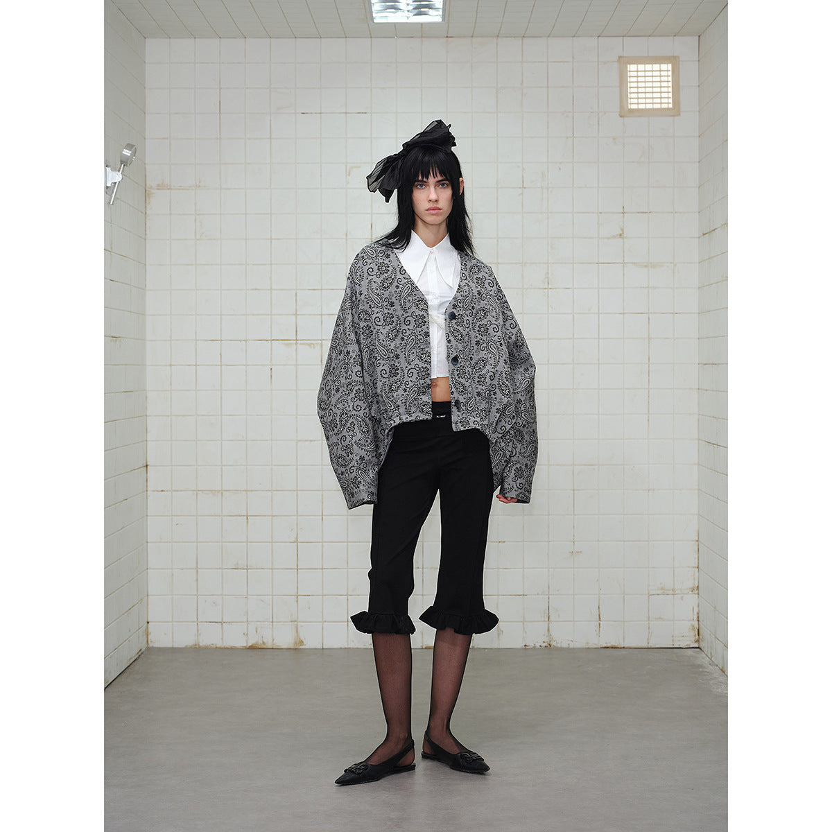 Front view of a model wearing an oversized gray paisley button-front jacket and black cropped pants with ruffled hems.