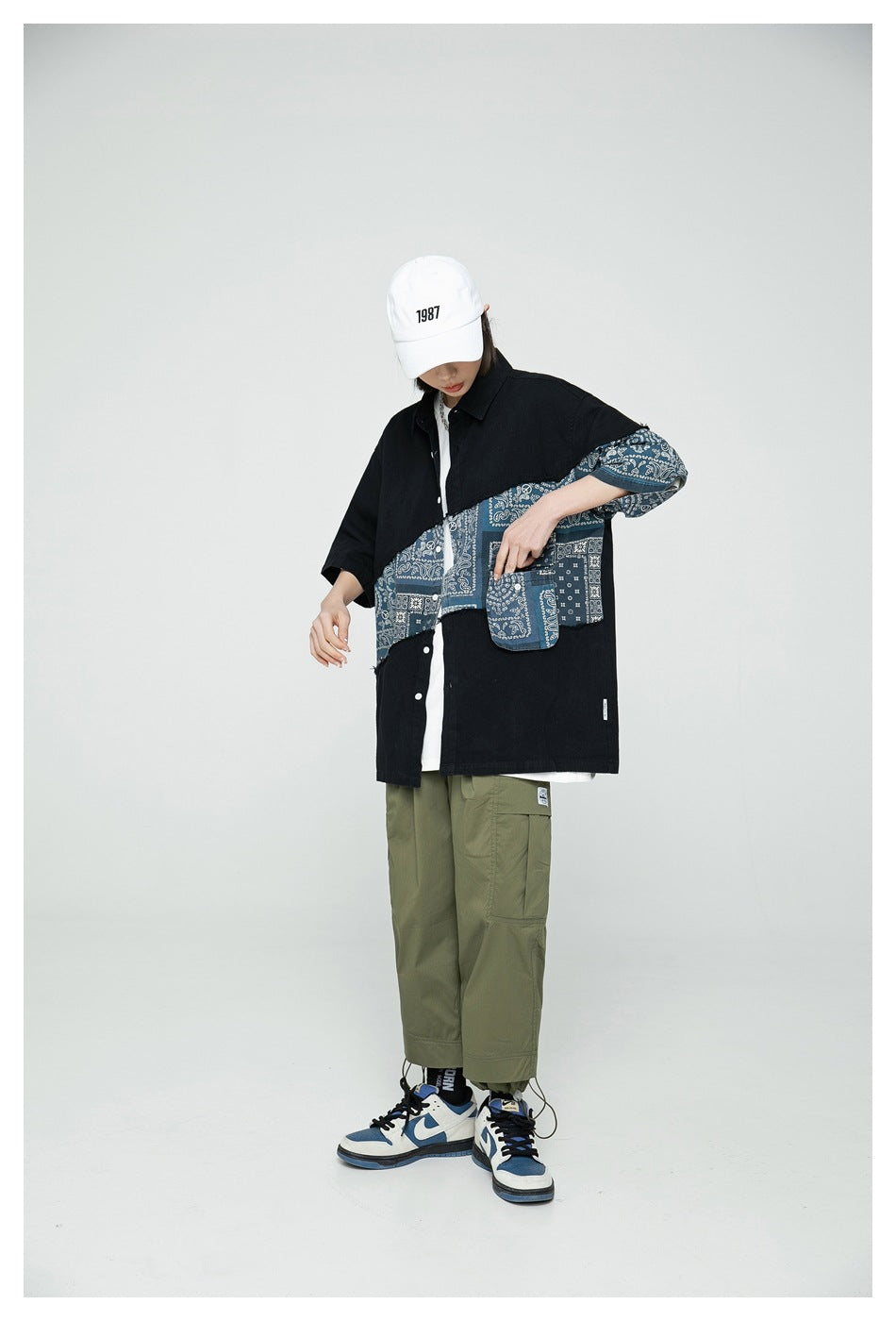 Model wearing black patchwork bandana shirt with flap pocket, white cap, olive cargo pants and blue and white sneakers