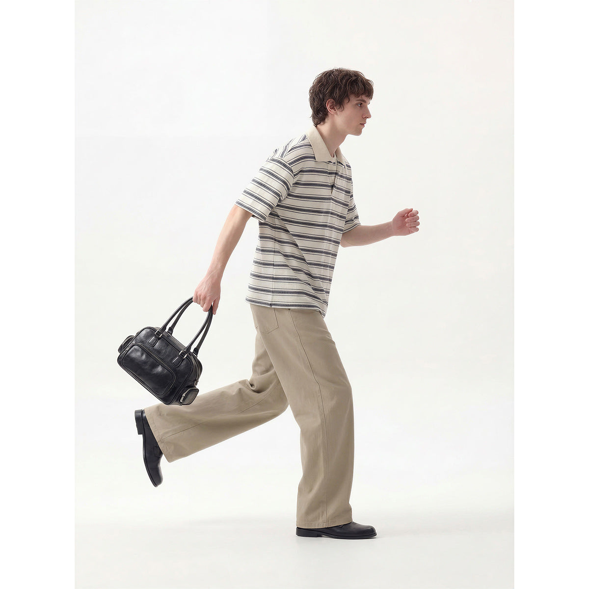 Model in a striped ivory polo and beige trousers, shown walking with a black bag.