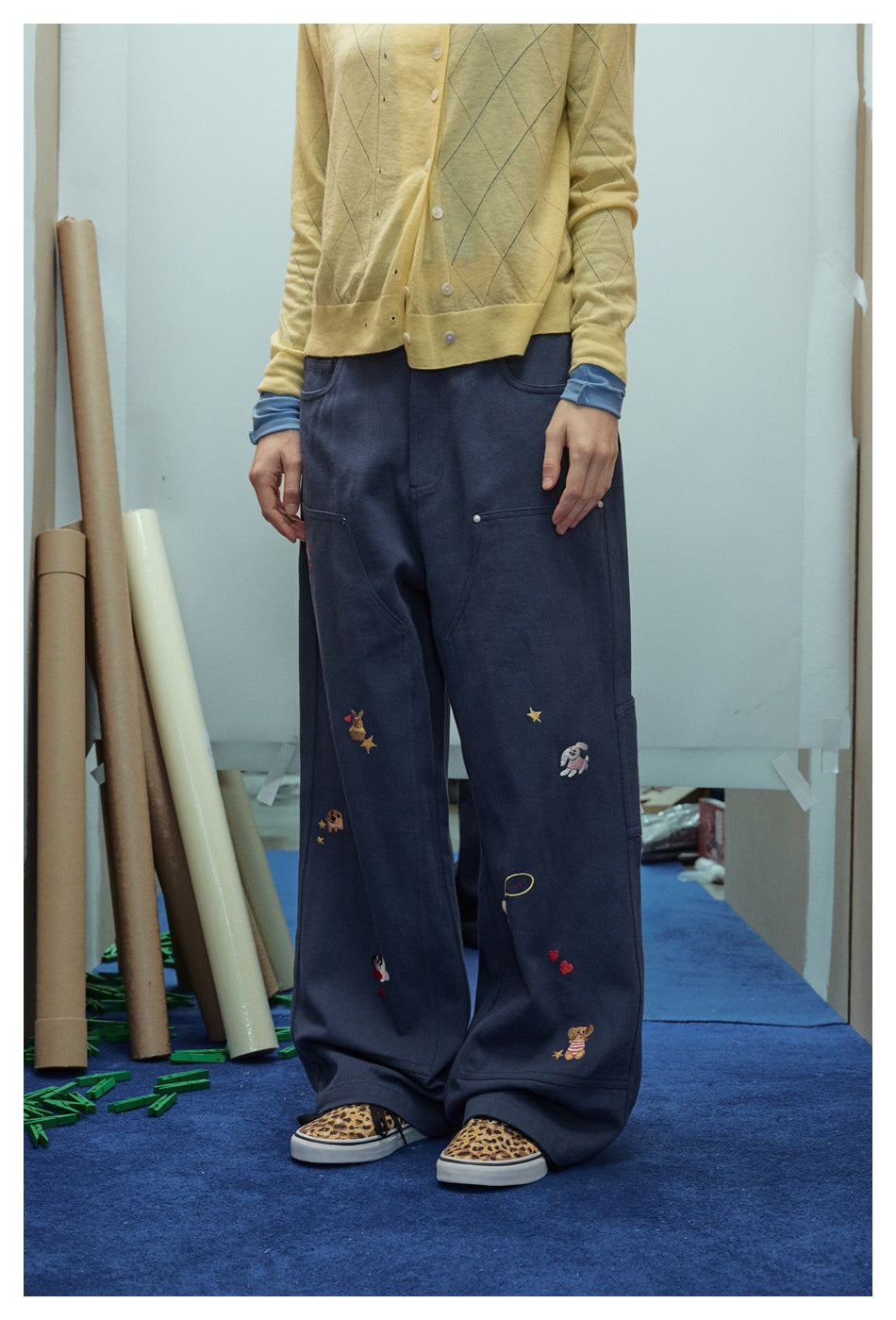 Person in navy wide-leg embroidered pants, yellow cardigan and leopard sneakers, angled front view cropped at the waist