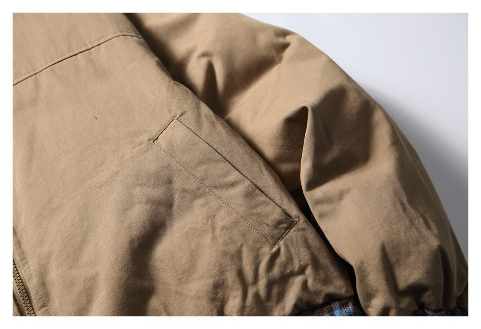 Close-up of a tan jacket’s slanted front pocket and sleeve, with plaid trim peeking at the hem.