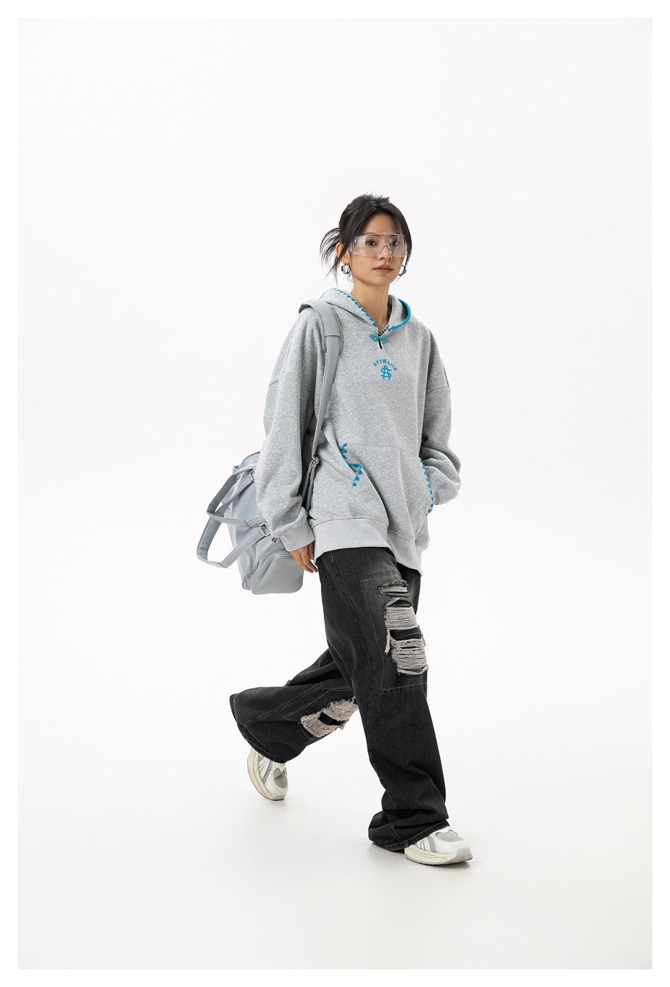 Full-length model wearing a gray hoodie with blue trim and black distressed jeans, carrying a gray bag.