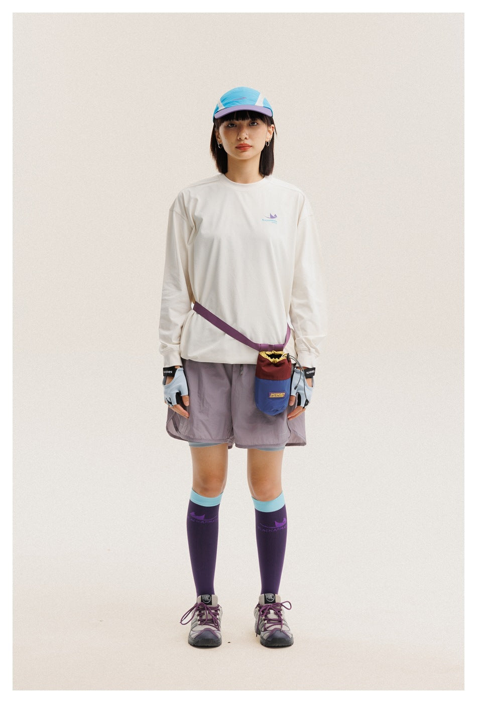 Front view of a woman wearing an off-white long-sleeve shirt and mauve shorts, with a cap, gloves, and a small crossbody