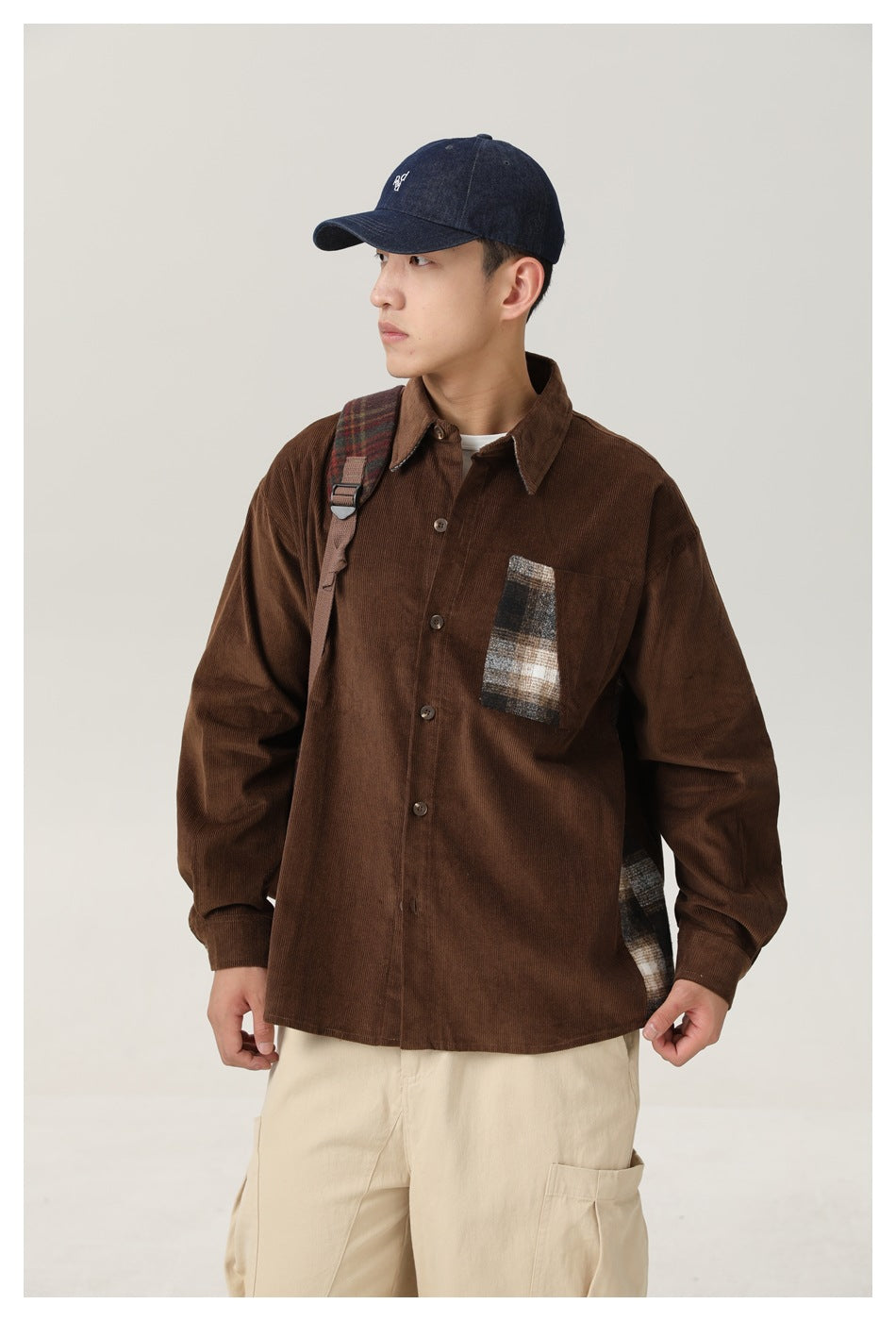 Closer front view of a model wearing a brown shirt with plaid chest and side accents, cream pants, and a cap.