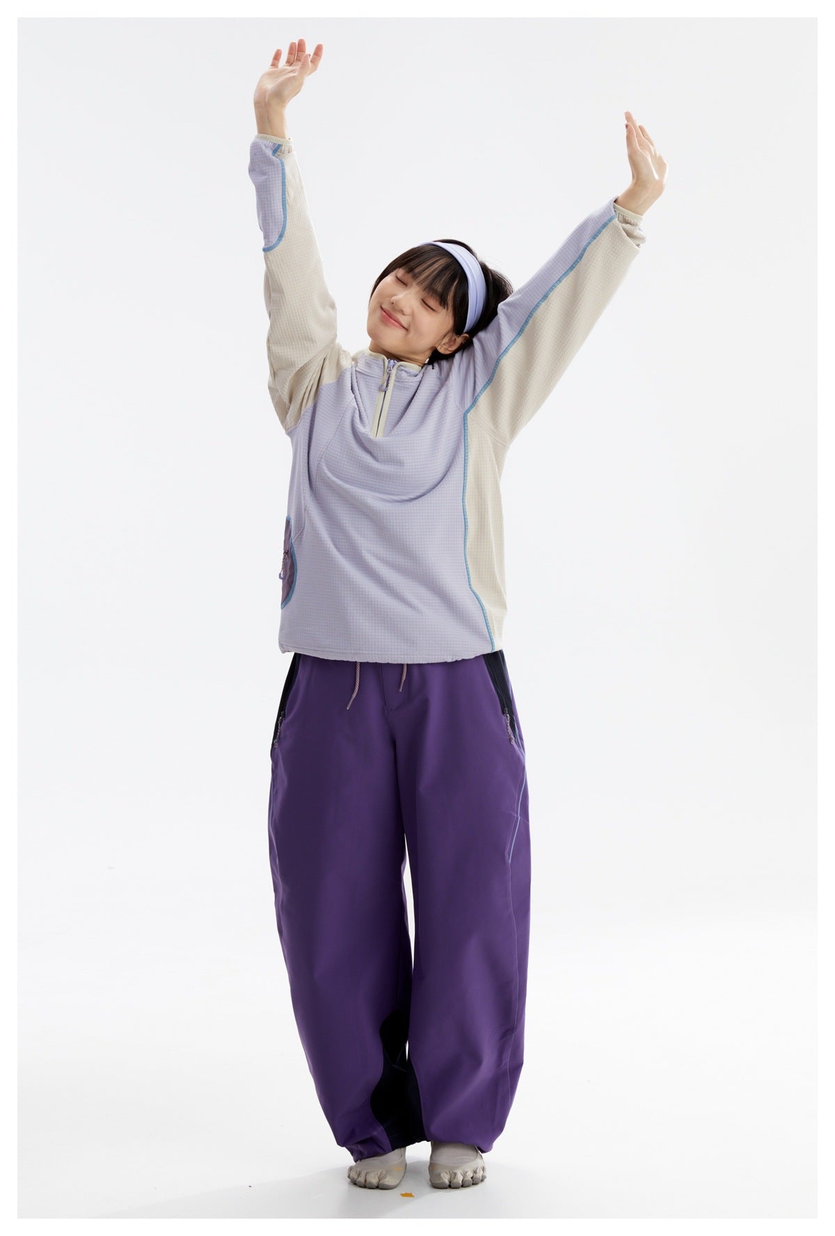 Model stretching in a lavender and cream color-block pullover with loose purple pants.