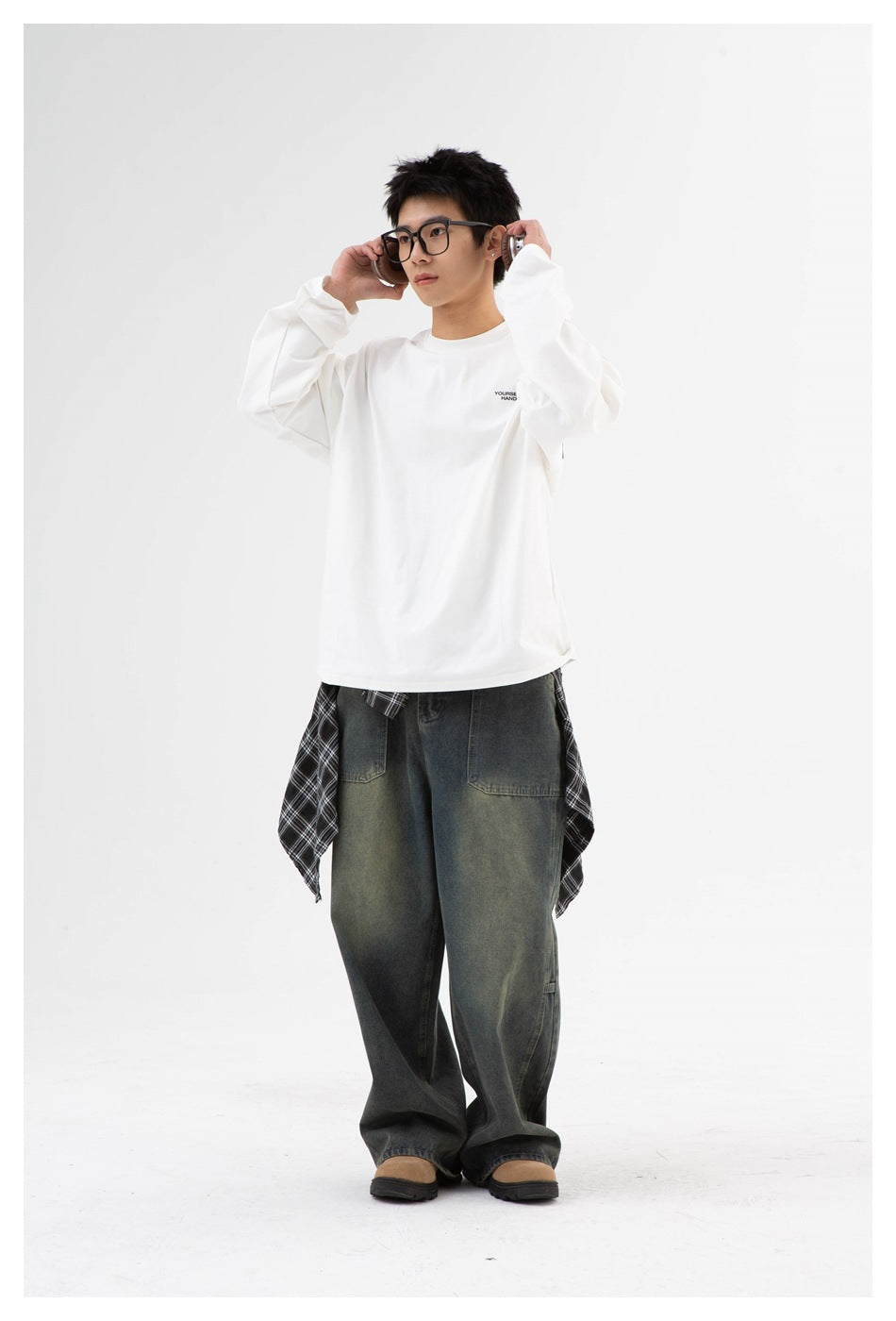 Model wearing a white oversized long-sleeve tee with small chest print, faded wide-leg jeans and a plaid shirt tied at the