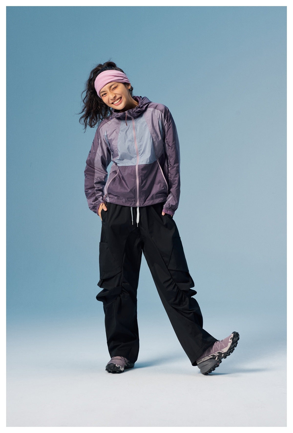 Model wearing a purple and pale blue color-block hooded jacket with black cargo pants, posing with one leg extended.