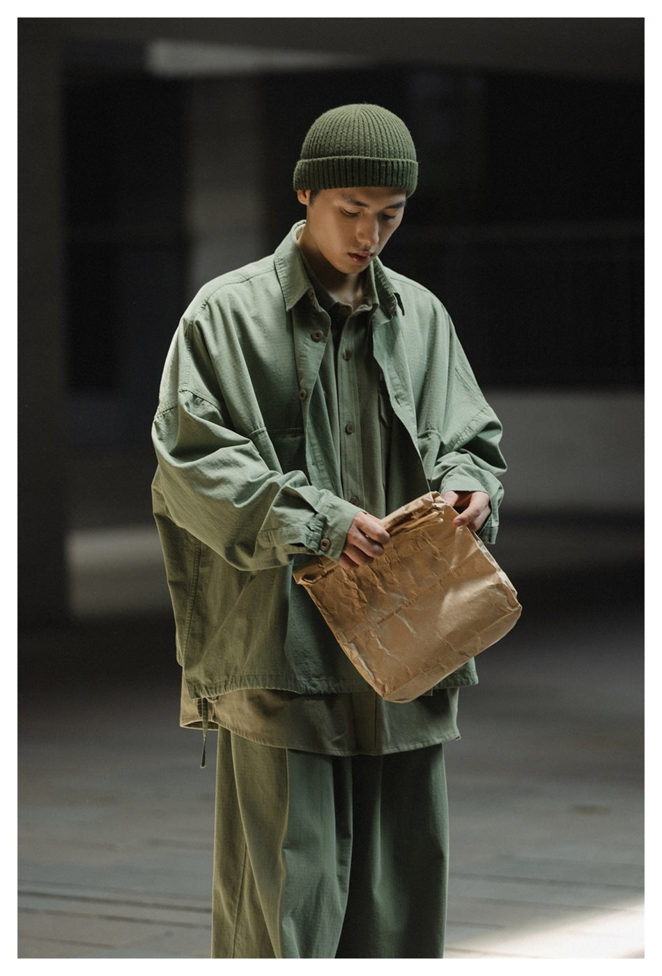 Man wearing an oversized army green shirt-jacket layered over a green shirt with a beanie, looking down at a paper bag