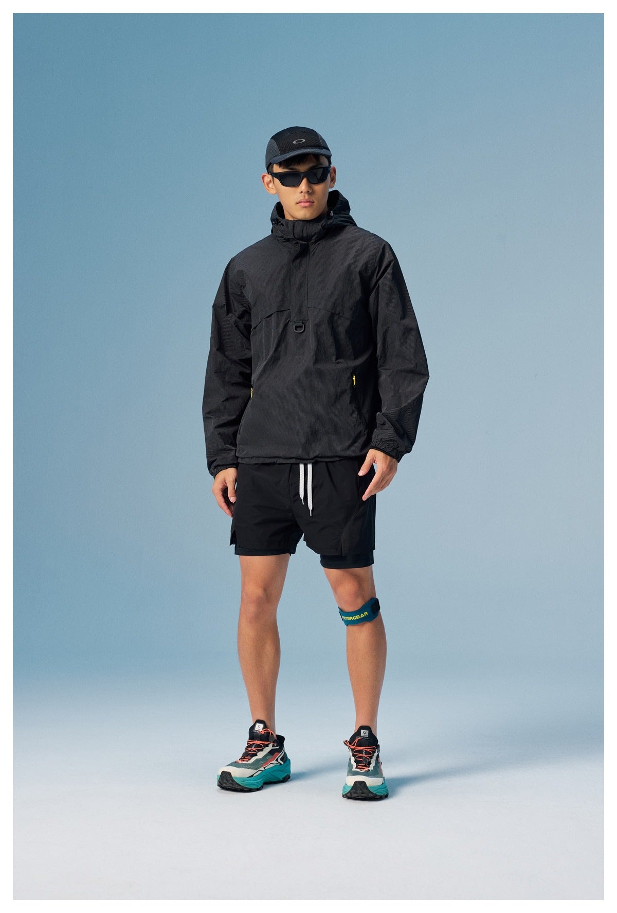 Model wearing a black hooded pullover and black shorts, with a cap, sunglasses, and running shoes.