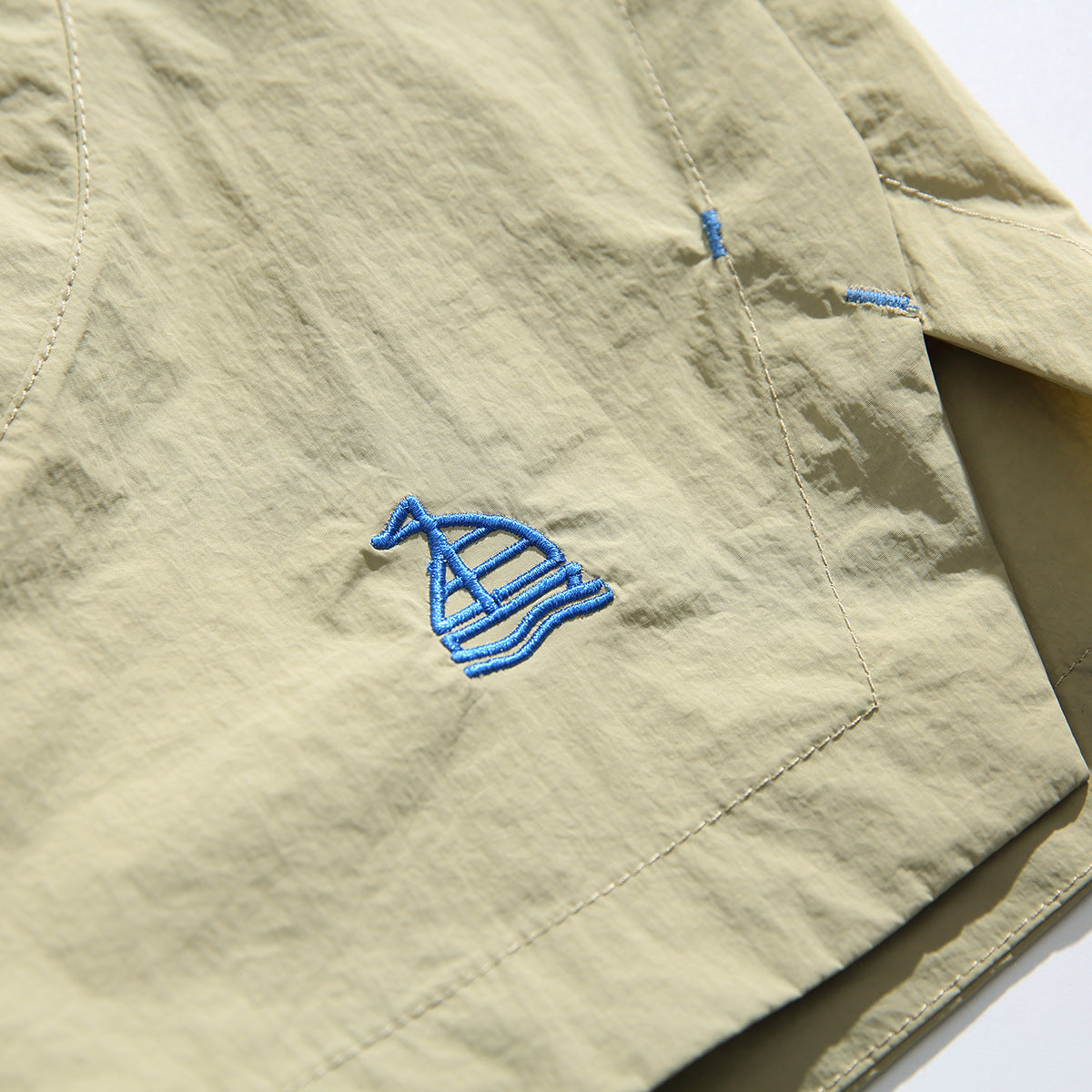 Close-up of beige shorts with a blue embroidered emblem, pocket stitching, and side seam.