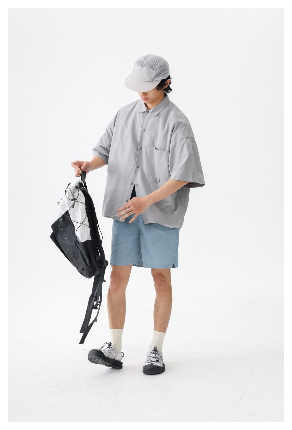 Model wearing a light gray short-sleeve button-up shirt and blue shorts, holding a black-and-white backpack.