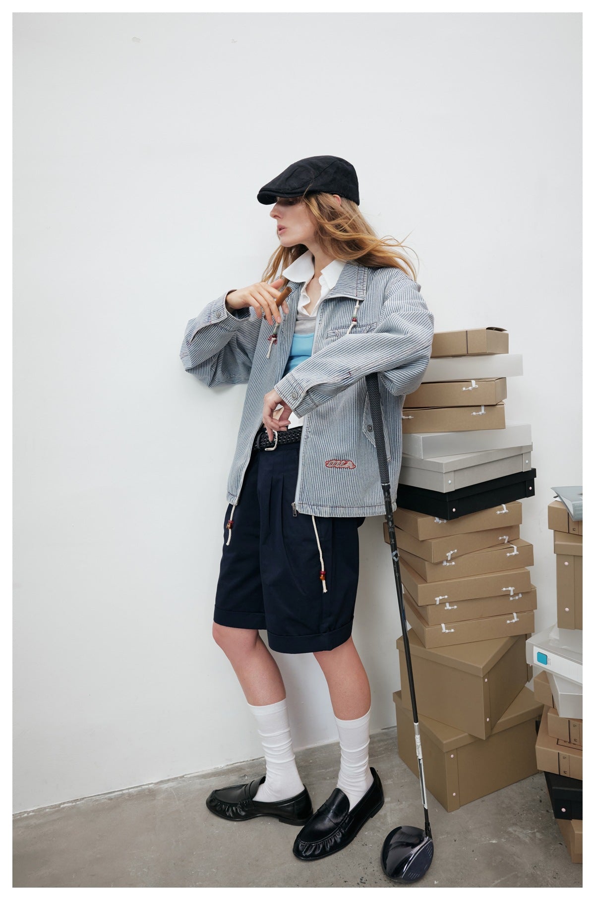 Model wearing a blue-and-white pinstriped jacket with navy shorts, a black cap, white socks, and black loafers.