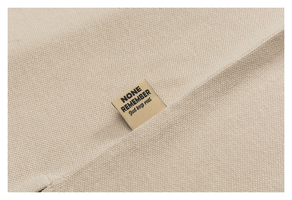 Close-up of a beige woven garment tag printed with “NONE REMEMBER just keep real.”