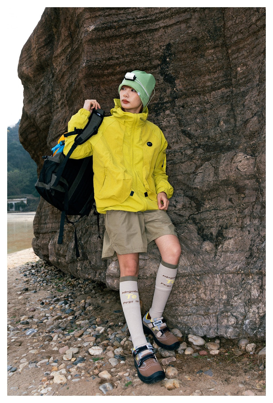 Person wearing a bright yellow hooded jacket, khaki shorts and green beanie, carrying a multicolor backpack outdoors.