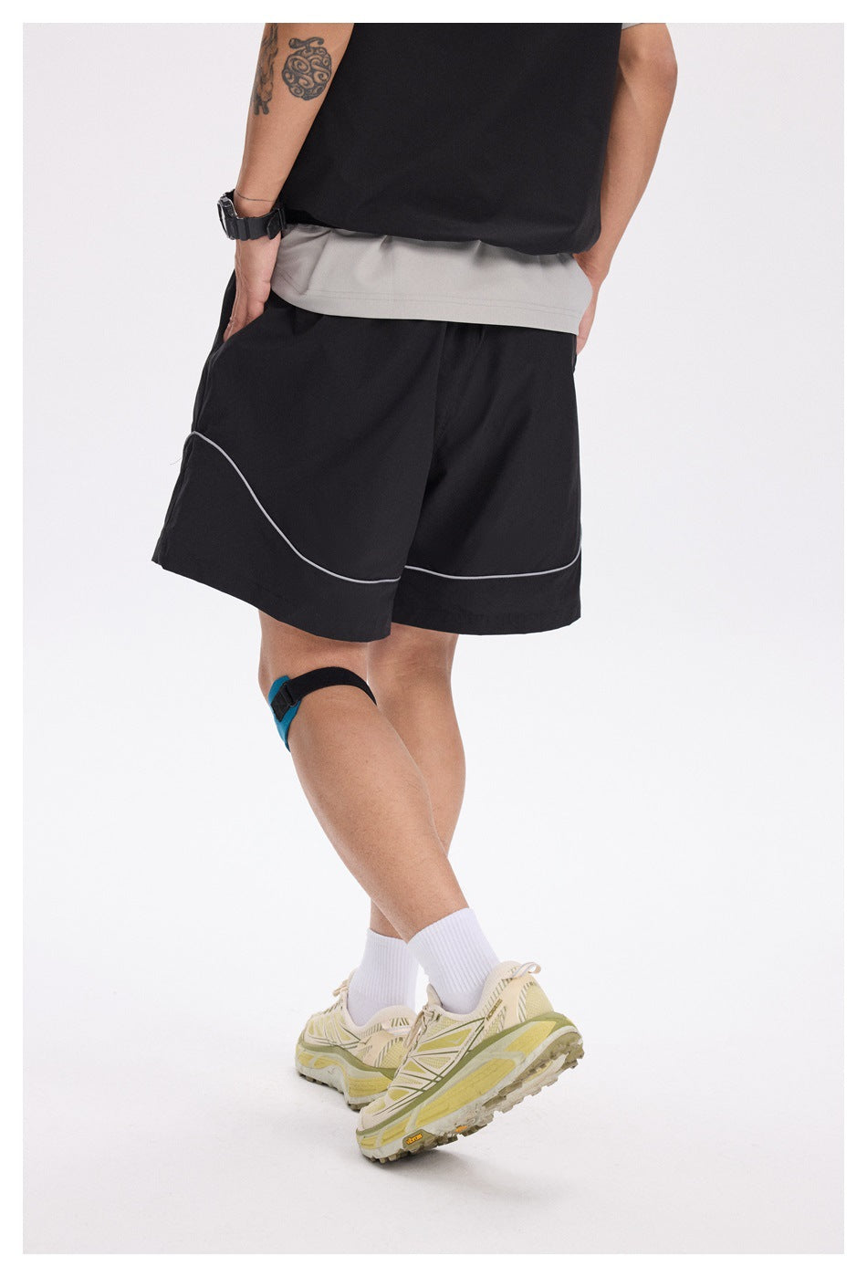 Rear view of a model wearing black shorts with curved gray piping, with a gray top and yellow sneakers