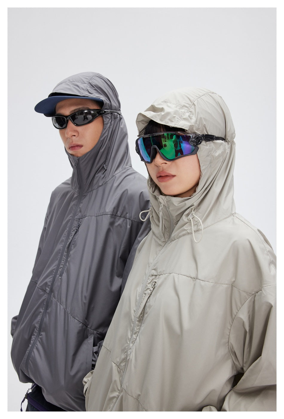 Close view of two models wearing gray and light beige hooded windbreakers with sunglasses.