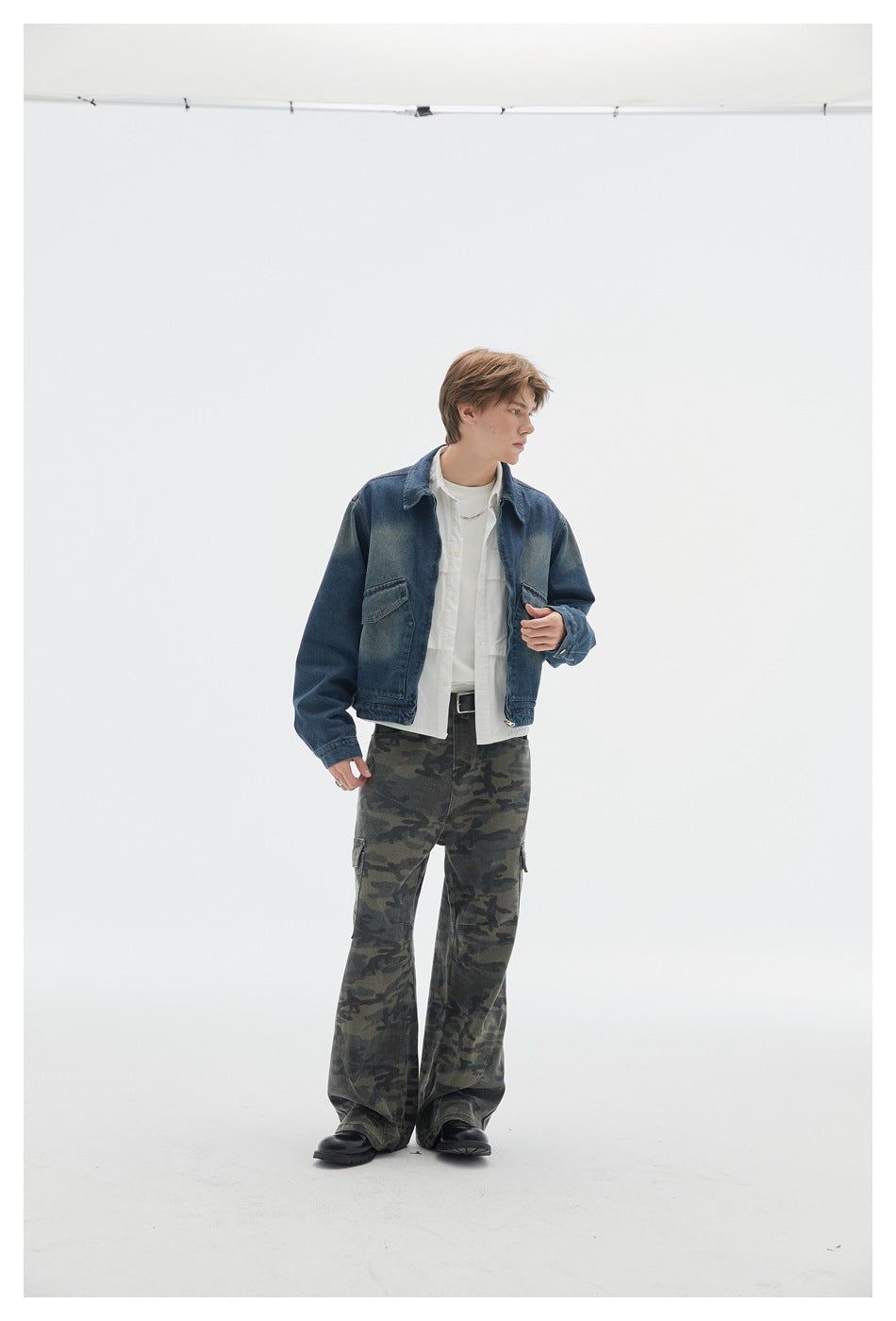 Model wearing a dark blue denim jacket, white layers, and loose camouflage cargo pants, turned slightly to the side.