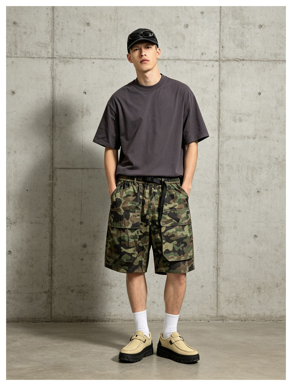 Model wearing camouflage cargo shorts with a gray tee, black cap, white socks and tan shoes against a concrete wall
