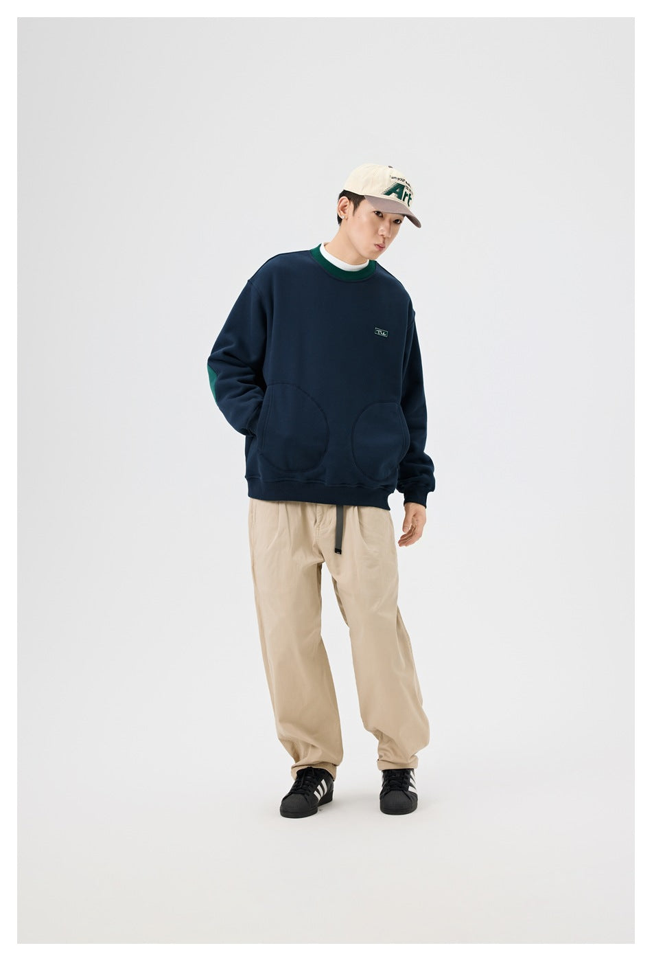 Model wearing a navy crewneck sweatshirt with green collar trim, elbow panels, and rounded front pockets.