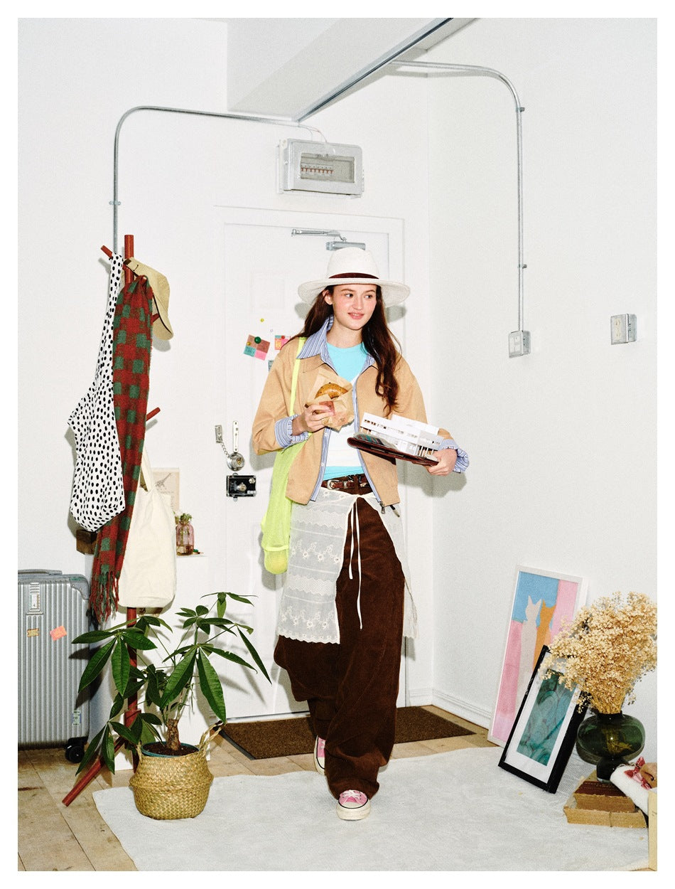 Woman walking in a khaki zip jacket with striped shirt layer, hat, brown trousers and lace skirt layer in a styled room