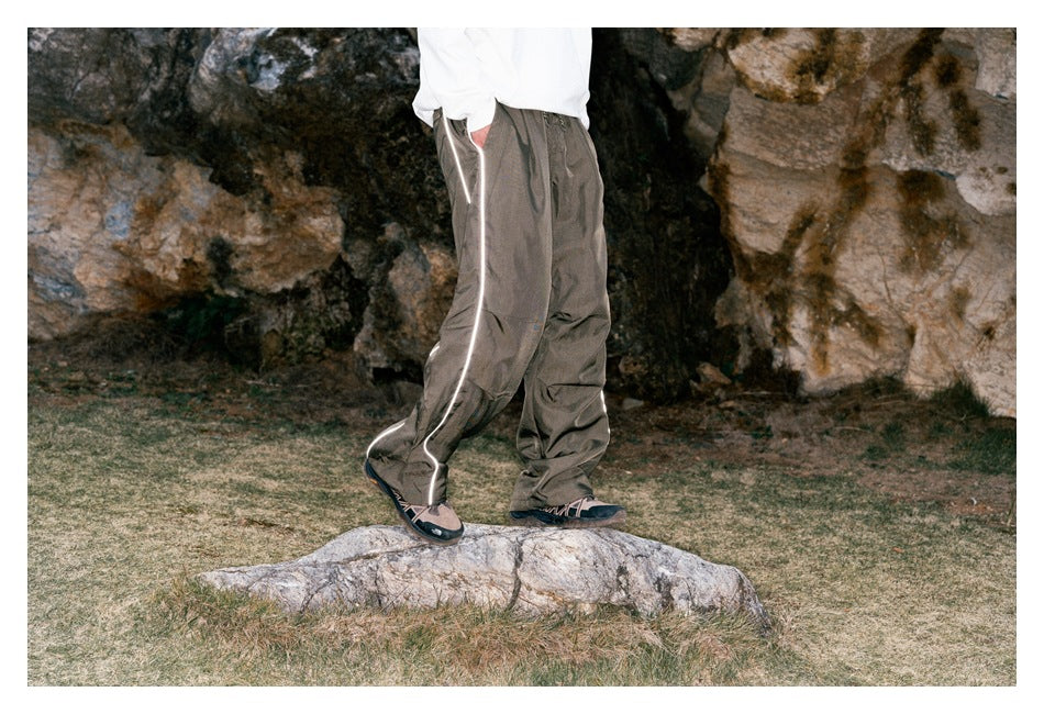 Three-quarter side view of loose taupe-gray pants with reflective piping, worn by a model outdoors.