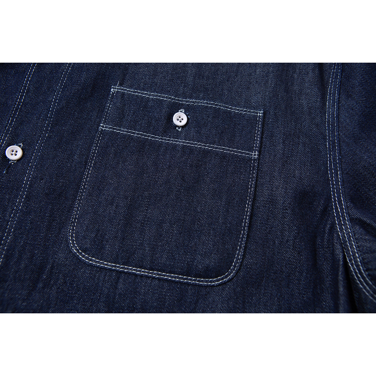 Close-up of a dark indigo shirt's rectangular chest pocket with white contrast stitching and a white button.