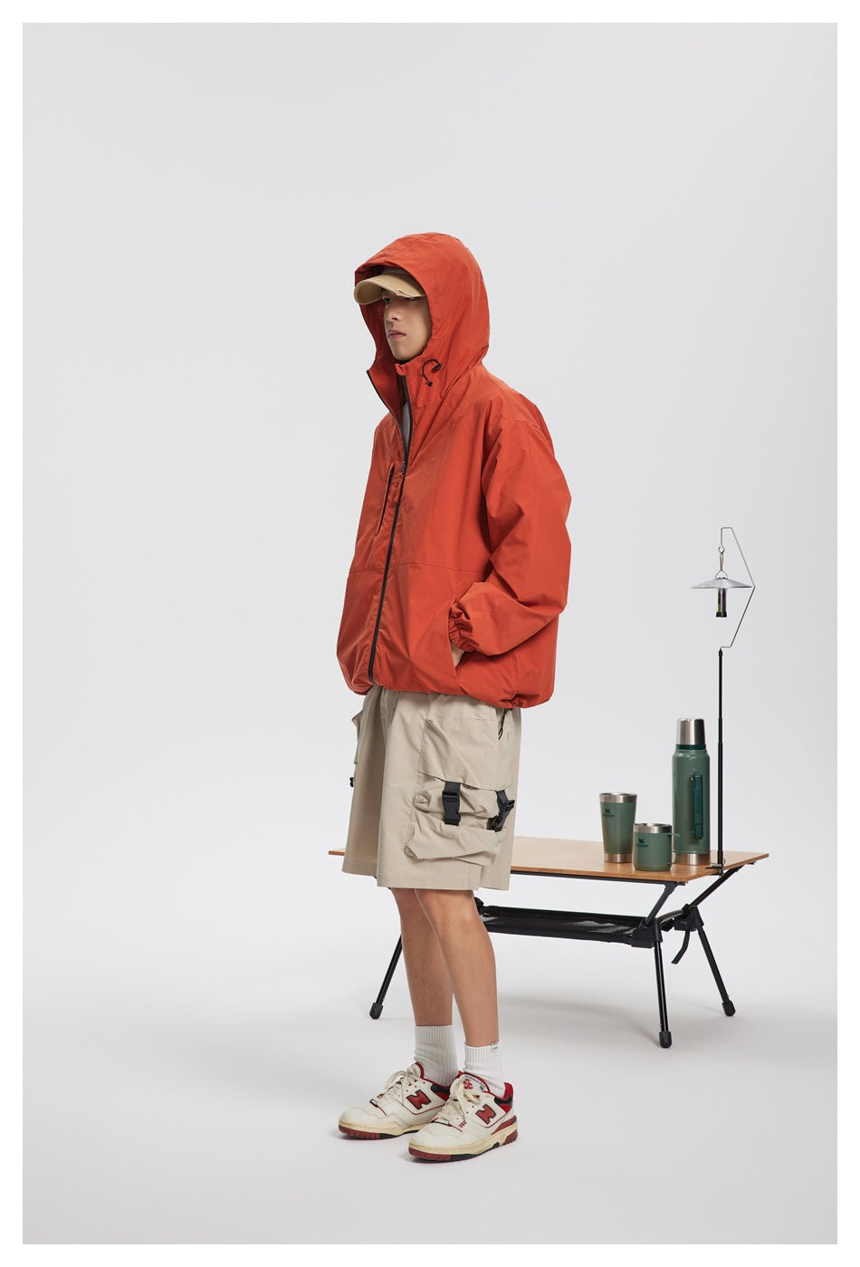 Full-length model wearing a rust-orange hooded jacket with the hood up and beige cargo shorts.