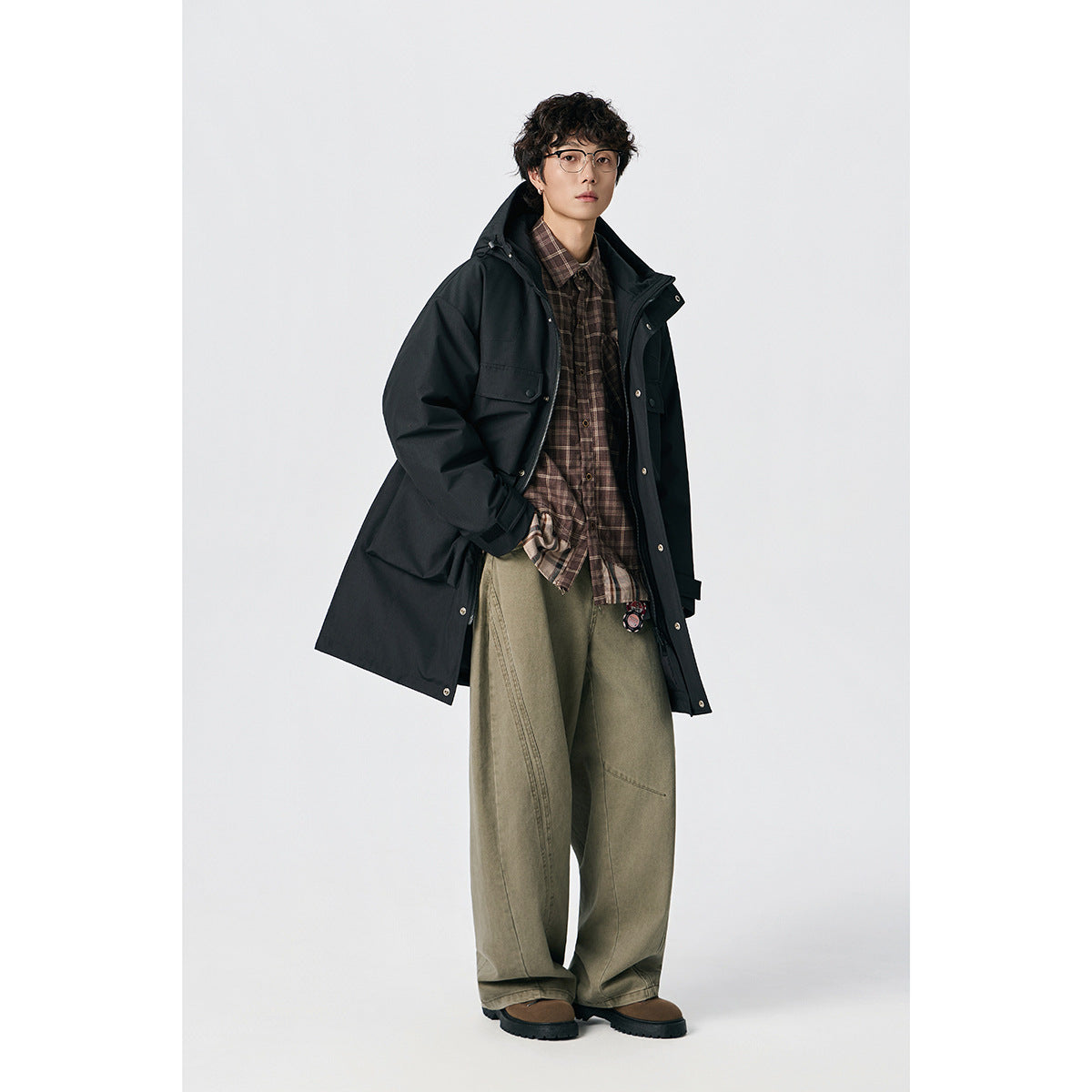 Model wearing an open black hooded long coat over a brown plaid shirt and loose khaki trousers.