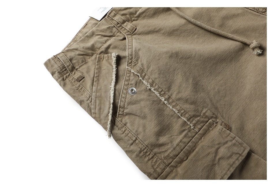 Close-up of taupe shorts showing a frayed pocket edge, metal snap, drawstring, and stitched seams.