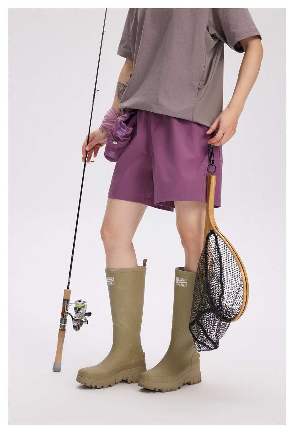 Person wearing plum-purple shorts with a gray shirt and khaki boots, holding fishing gear; front view.